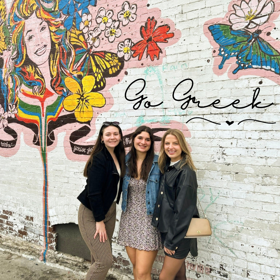 three girls pose in front of a mural, smiling at the camera. Above them are the words go greek.