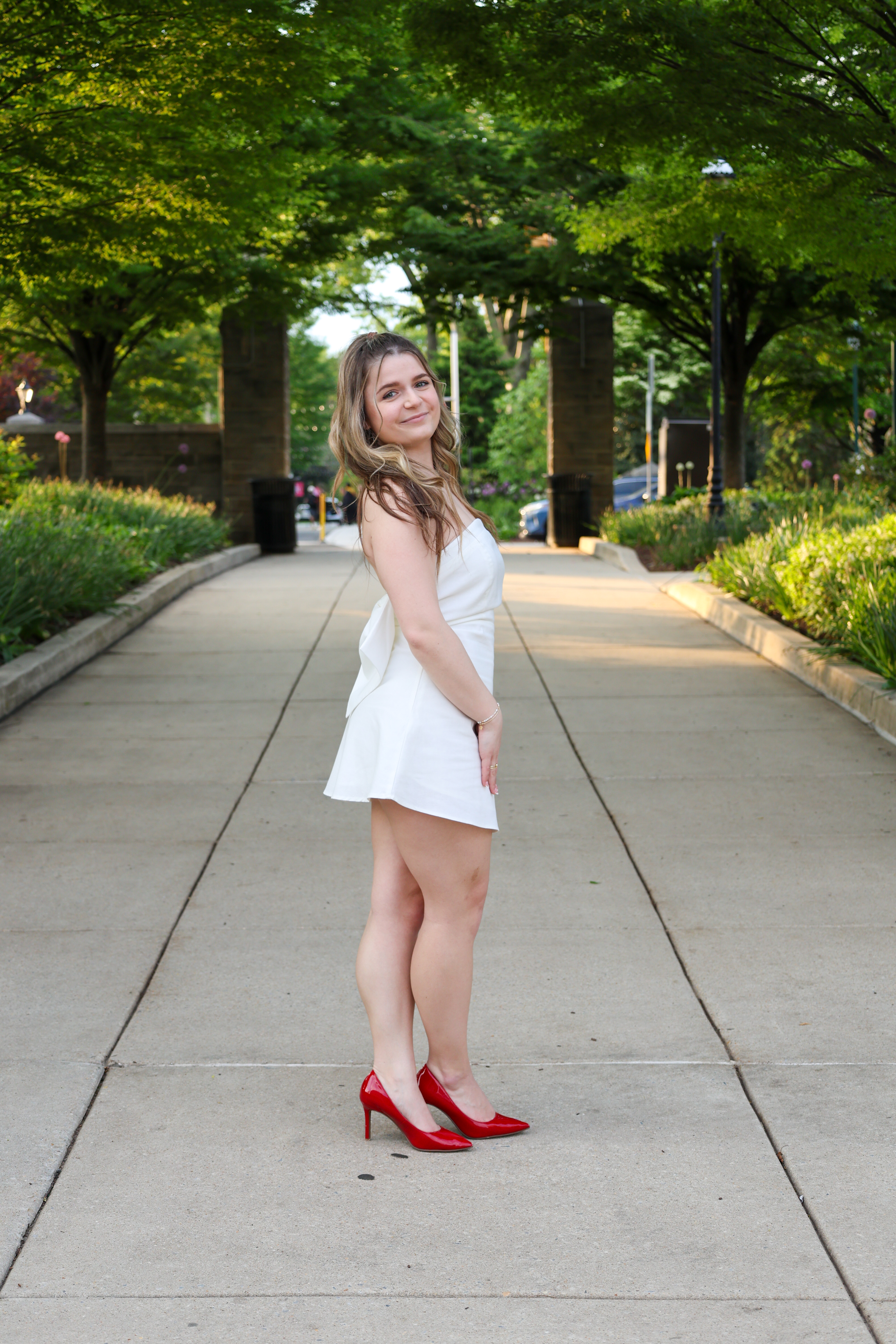 Color photo. Subject is standing on Saint Joseph's campus in a white graduation dress, smiling at camera.