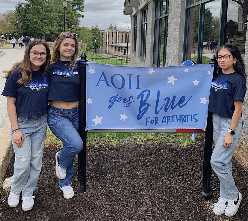 picture of Kaylie and two other girls wearing navy blue shirts with the design on it. They are standing in front of a sign that says AO Pi goes blue for arthritis.