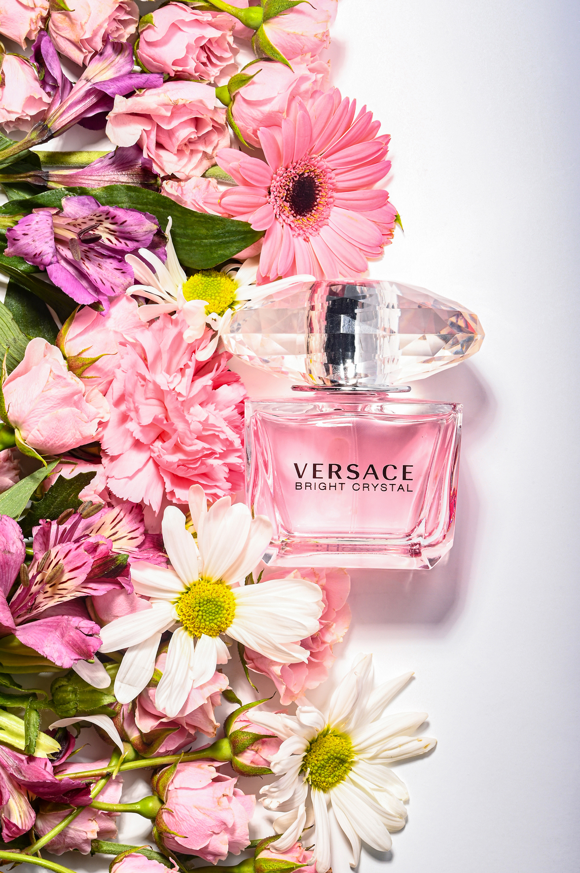 Color photo. Versace perfume bottle with pink liquid, surrounded by an assortment of flowers..